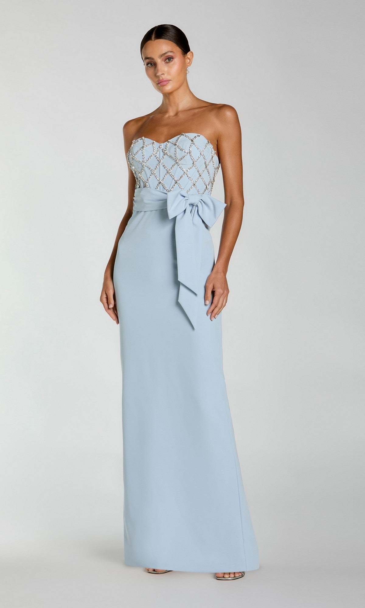 A woman models the Mac Duggal 2294 Strapless Sweetheart Formal Gown with a fitted lattice bodice and large waist bow, standing against a plain background.